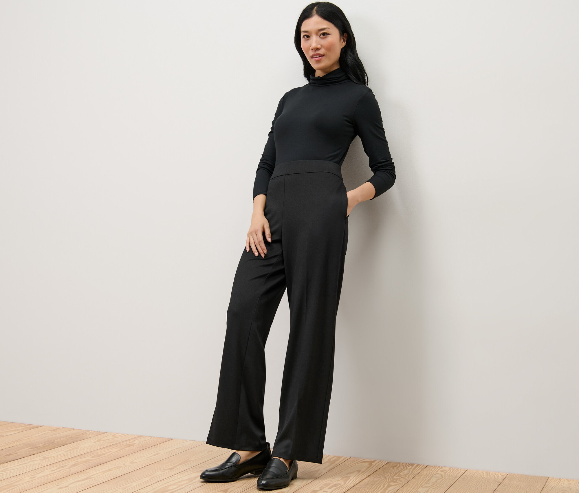 Žena v černém roláku a pohodlných žerzejových kalhotách stojí opřená o zeď.