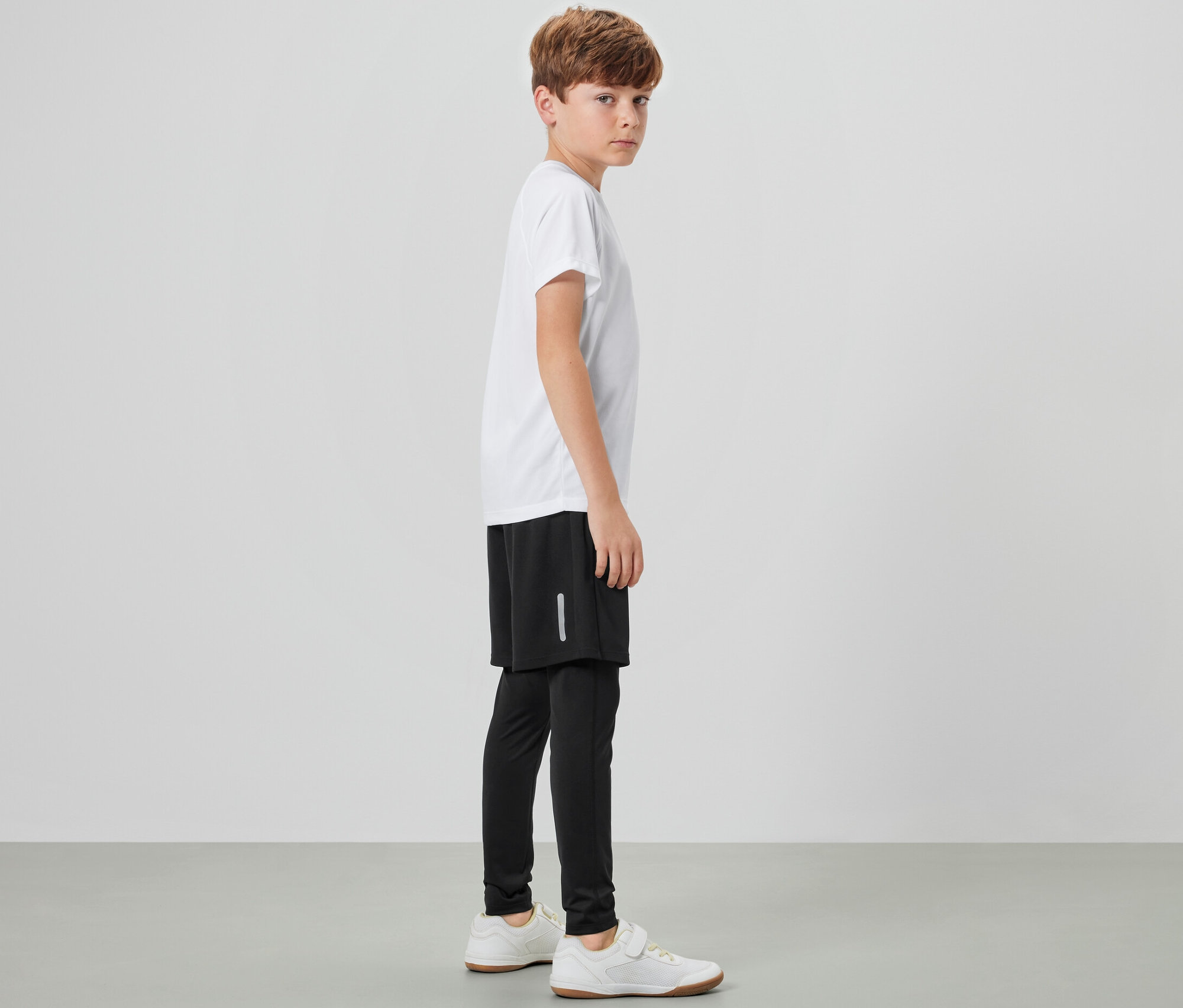 Chlapec stojí bokem a má na sobě dětské sportovní legíny s šortkami, bílé tričko a tenisky.