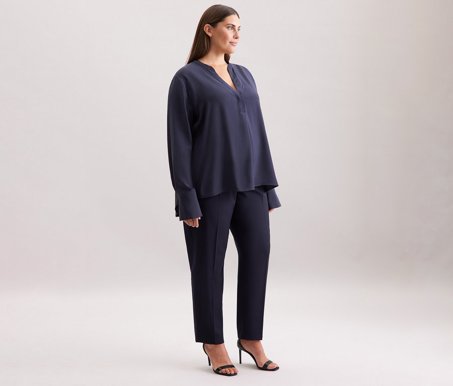 Žena stojí bokem, má na sobě námořnicky modrou tuniku Seidensticker z viskózy, kalhoty a sandály.