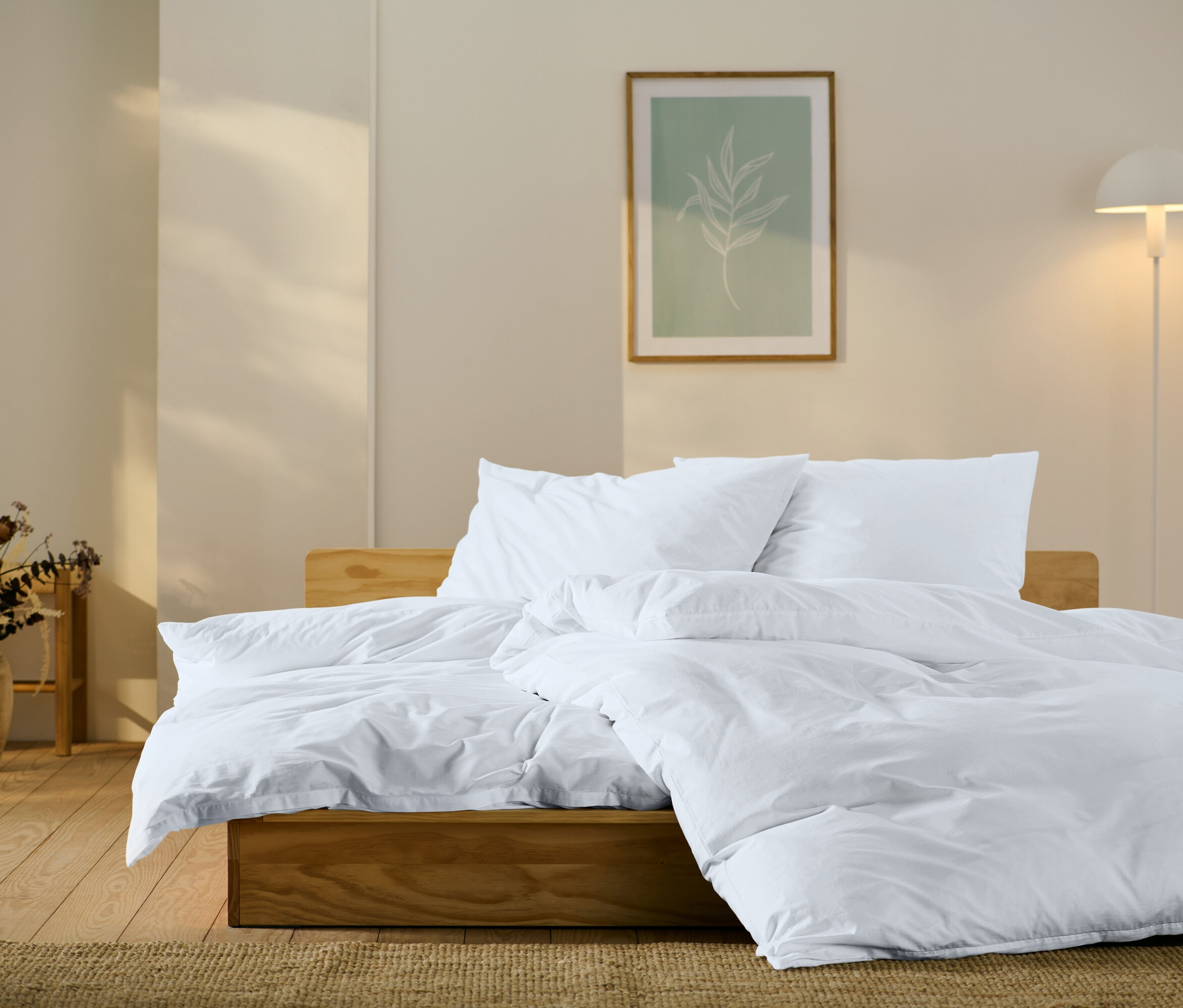 Dřevěná postel s bílým povlečením, polštáři a přikrývkou. Na zdi obraz s motivem listu a lampa.