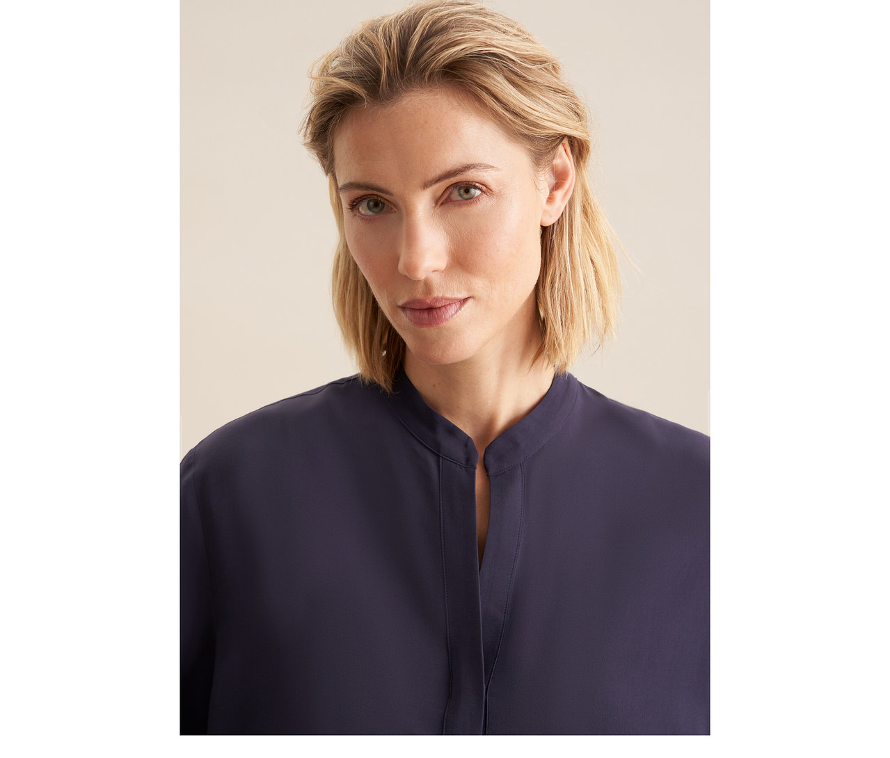 Žena s blond vlasy má na sobě tmavomodrou tuniku z viskózy Seidensticker se stojáčkem a výstřihem do V.