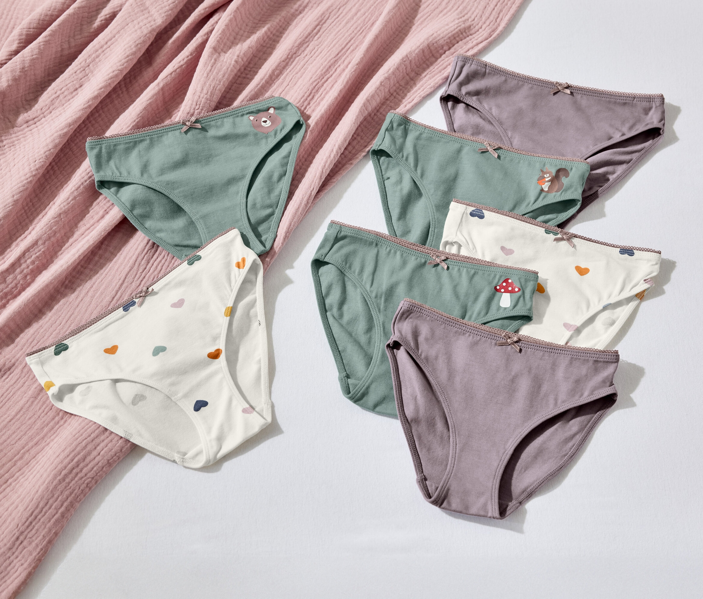 Sedm párů dámských kalhotek různých barev a vzorů leží na bílém pozadí vedle růžové látky.