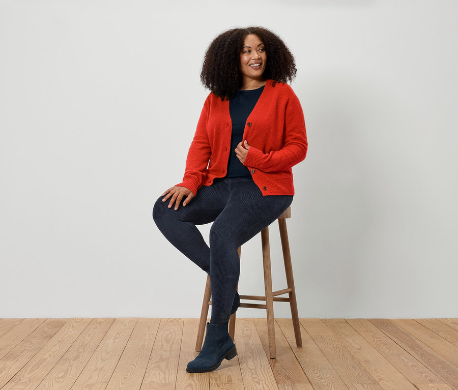 Žena sedí na stoličce a má na sobě červený kardigan, modré džíny a modré boty.