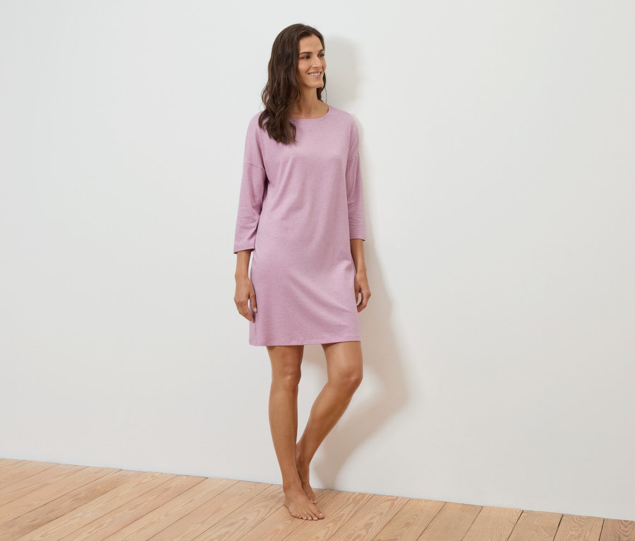 Žena stojí a usmívá se, má na sobě fialové šaty s tříčtvrtečními rukávy.