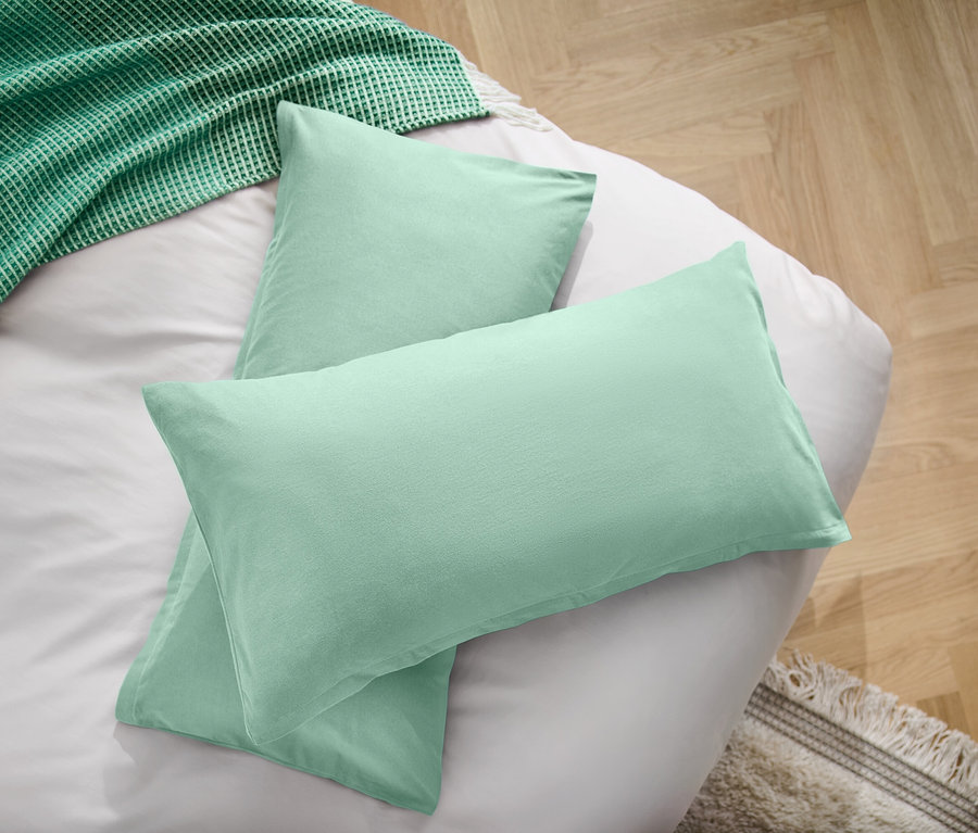 Dva světle zelené polštáře na bílém prostěradle vedle zelené deky.