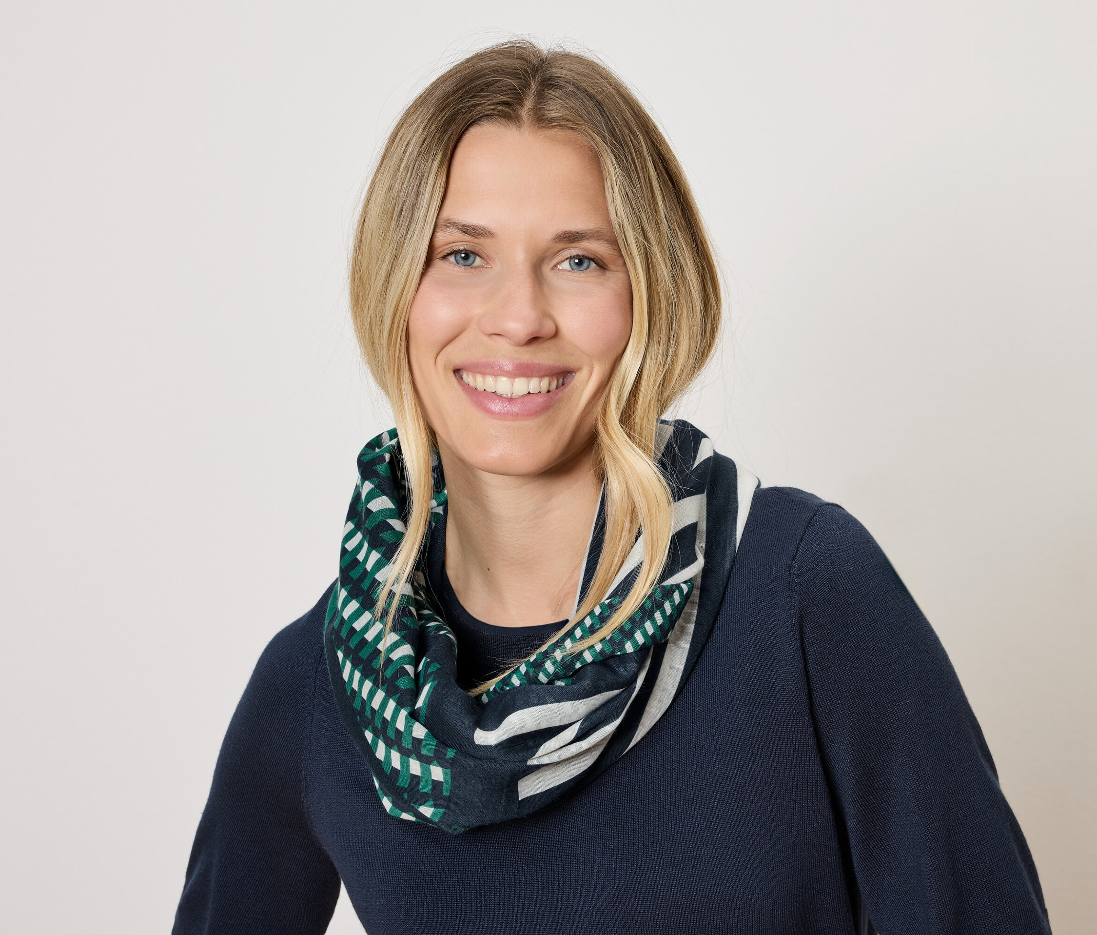Žena s blond vlasy a úsměvem má na sobě tmavě modrý svetr a šátek s geometrickým vzorem v zelené a bílé barvě.