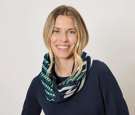 Žena s blond vlasy a úsměvem má na sobě tmavě modrý svetr a šátek s geometrickým vzorem v zelené a bílé barvě.