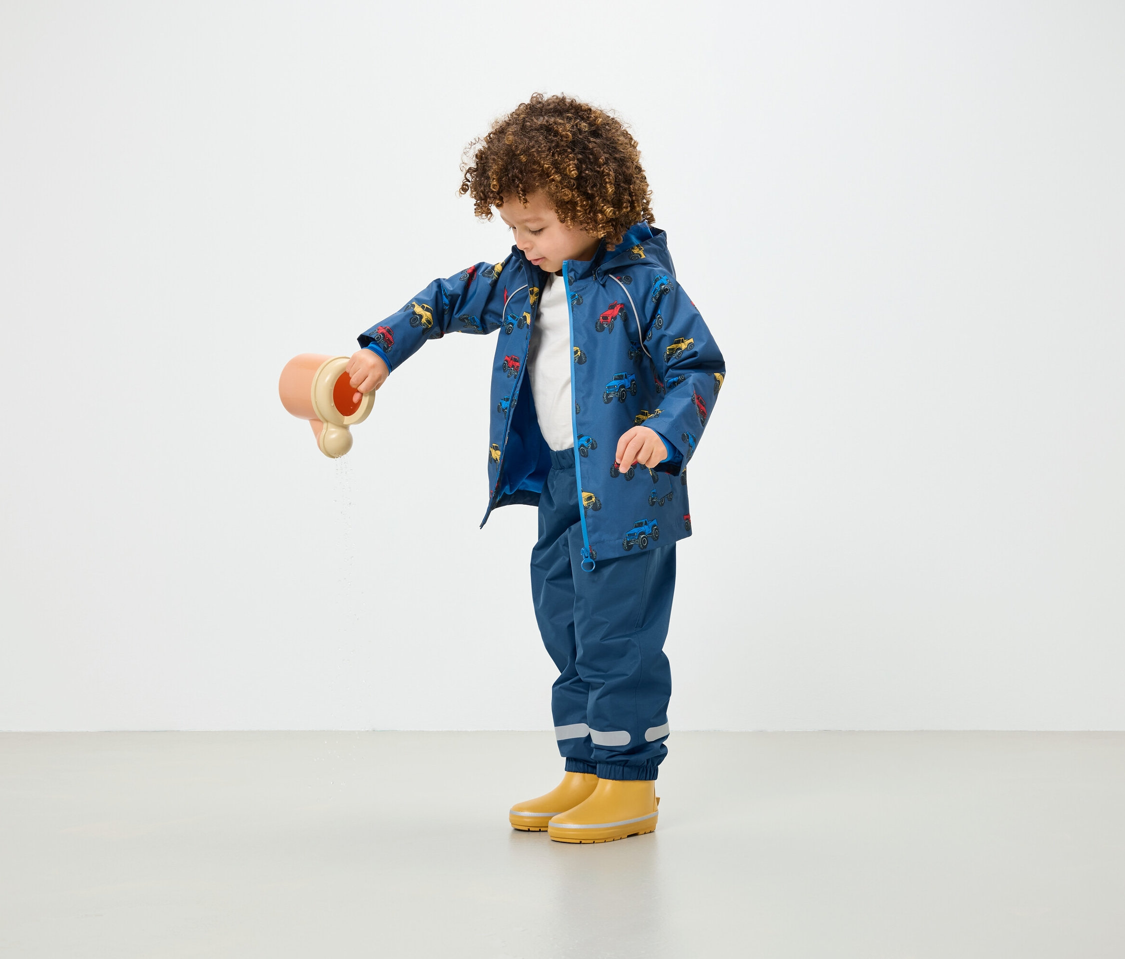 Dítě s kudrnatými vlasy nalévá vodu z hračky v modrém kabátě s potiskem a modrých kalhotách.