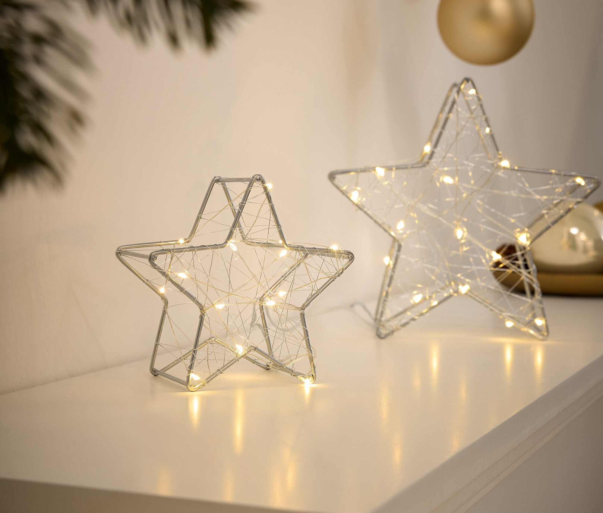 Dvě stříbrné drátěné hvězdy s malými světýlky stojí na bílém povrchu.