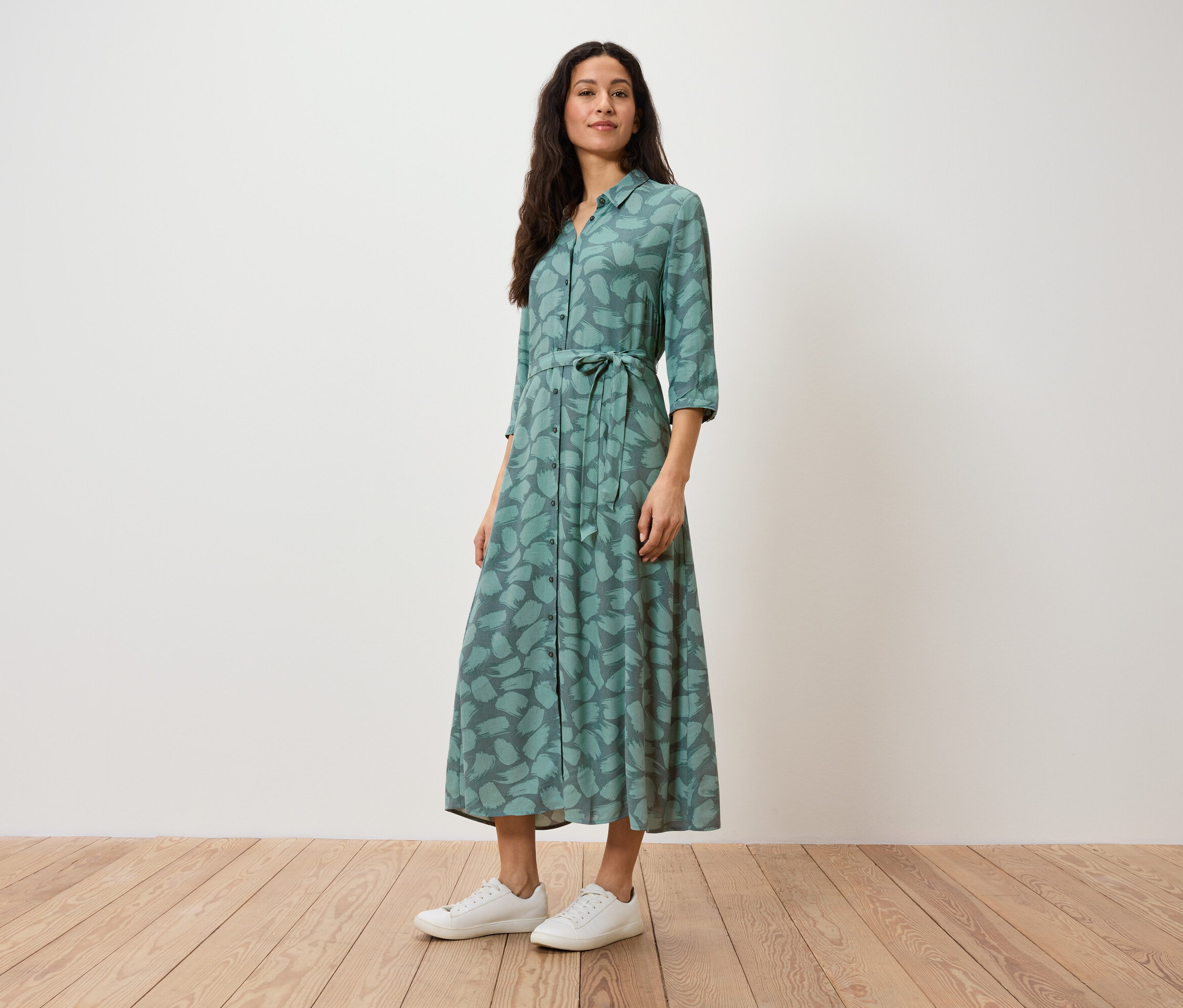 Žena stojí a má na sobě zelené košilové šaty s potiskem a bílé tenisky.