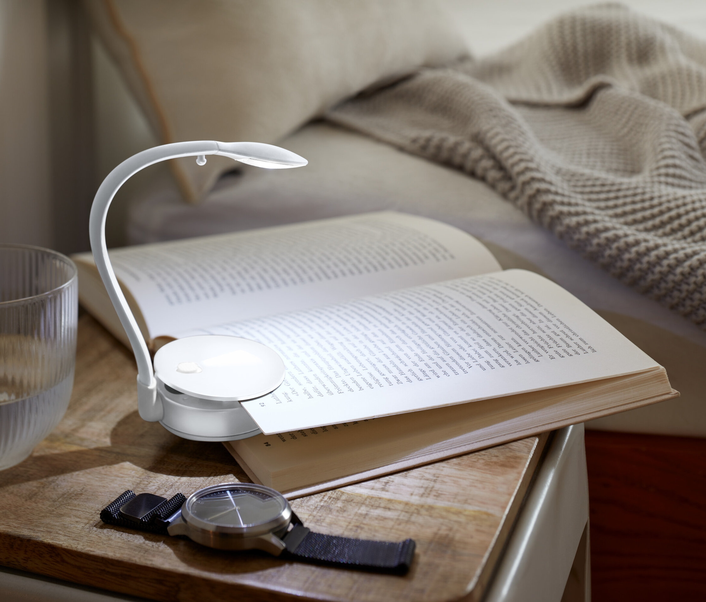 Na nočním stolku leží otevřená kniha osvětlená malou lampičkou, vedle ní stojí sklenice s vodou a hodinky.