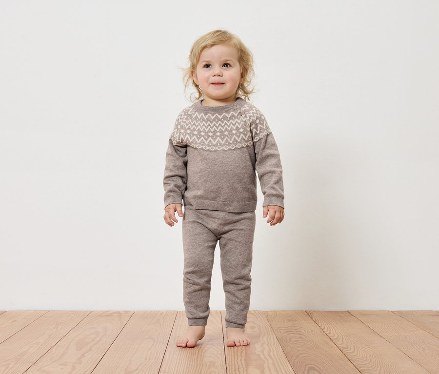 Batole stojí na dřevěné podlaze a má na sobě hnědý svetr a kalhoty.