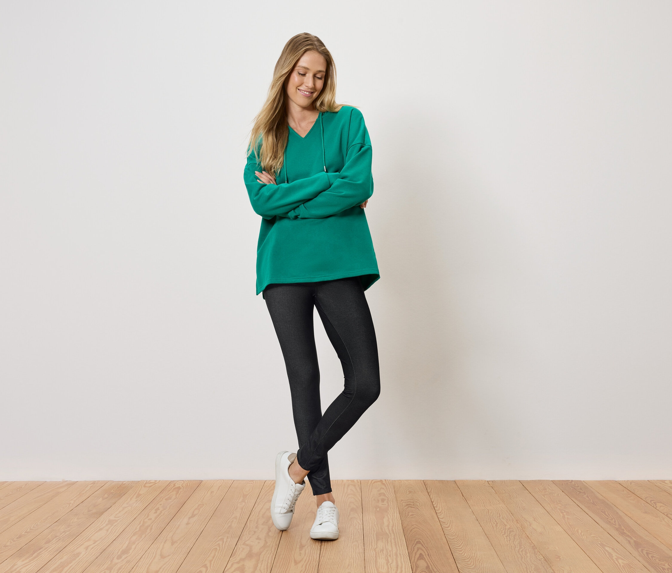 Žena stojí s rukama překříženýma na hrudi a usmívá se, oblečená do zeleného svetru, černých legín a bílých bot.
