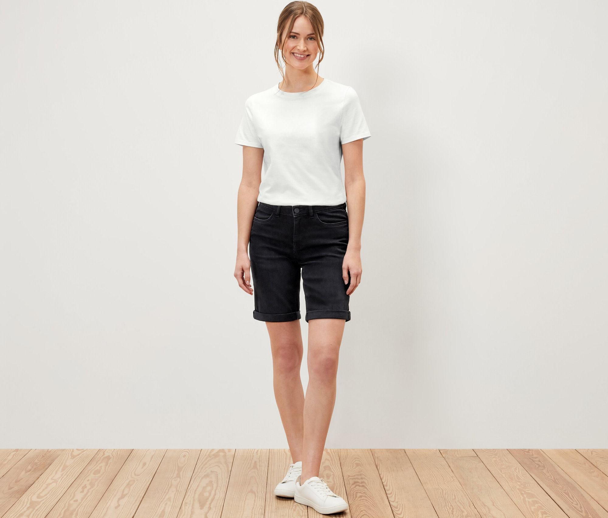 Žena stojící na dřevěné podlaze má na sobě džínové bermudy »Lea« černý denim a bílé tričko.