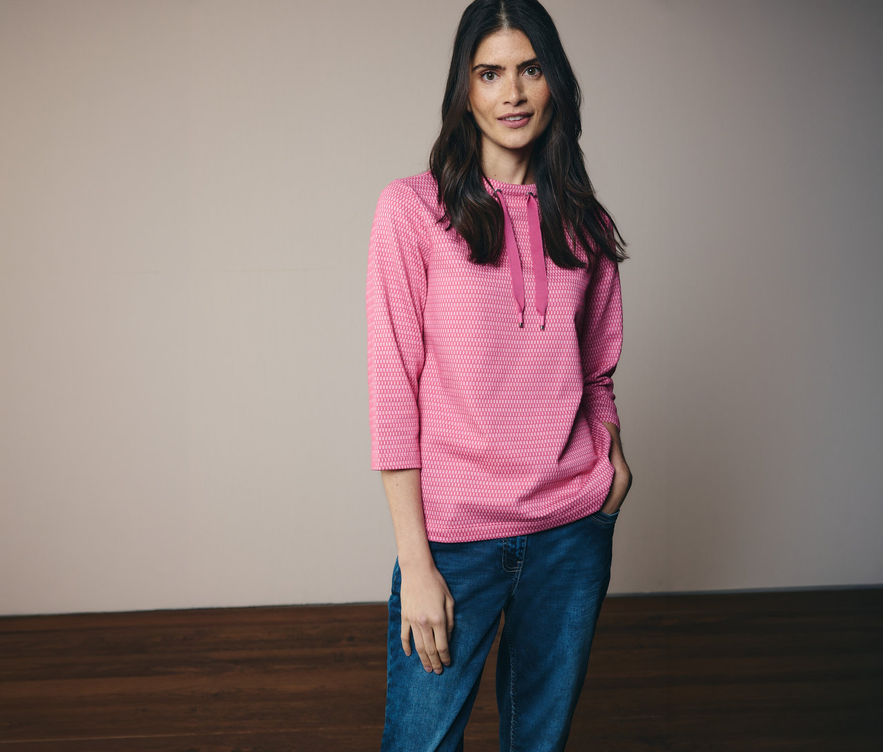 Žena v džínách a růžové mikině CECIL s kapucí a 3/4 rukávy, s rukou v kapse.
