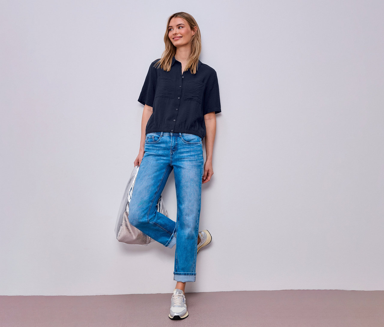 Žena v džínách a košilové halence Street One pózuje se stříbrnou taškou u zdi.