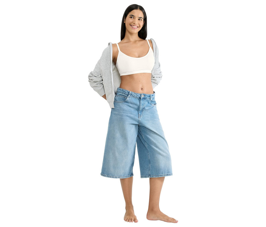Žena stojí bosá, má na sobě bílý crop top, široké džínové šortky a šedou bundu.