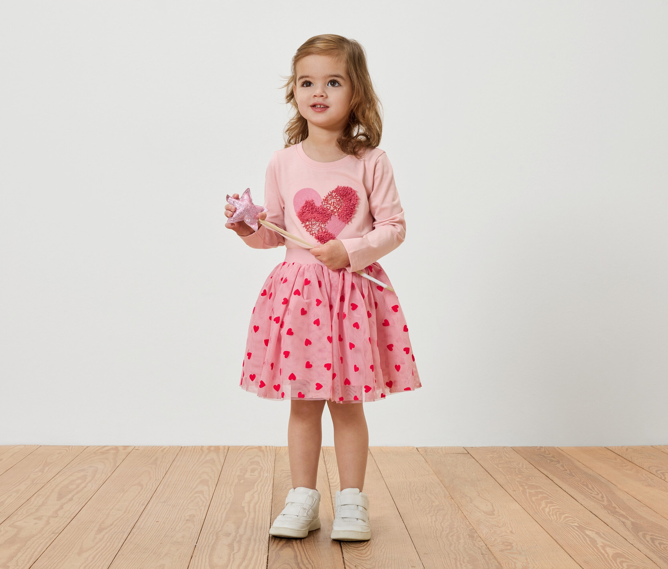 Malá blondýnka má na sobě růžové šaty se srdíčky a drží hůlku.