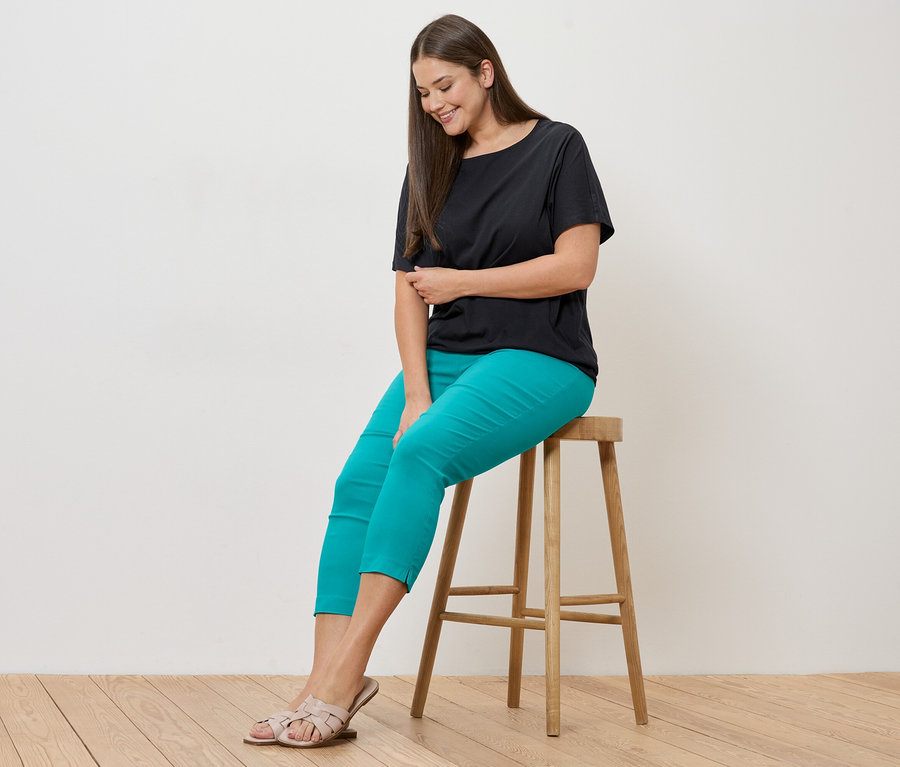 Žena sedí na stoličce, má na sobě černé blusenshirt a azurově modré 7/8-Stretchhose.