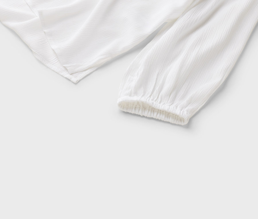 Detailní pohled na bílou halenku s dlouhými rukávy a nařasenými manžetami na bílém pozadí.