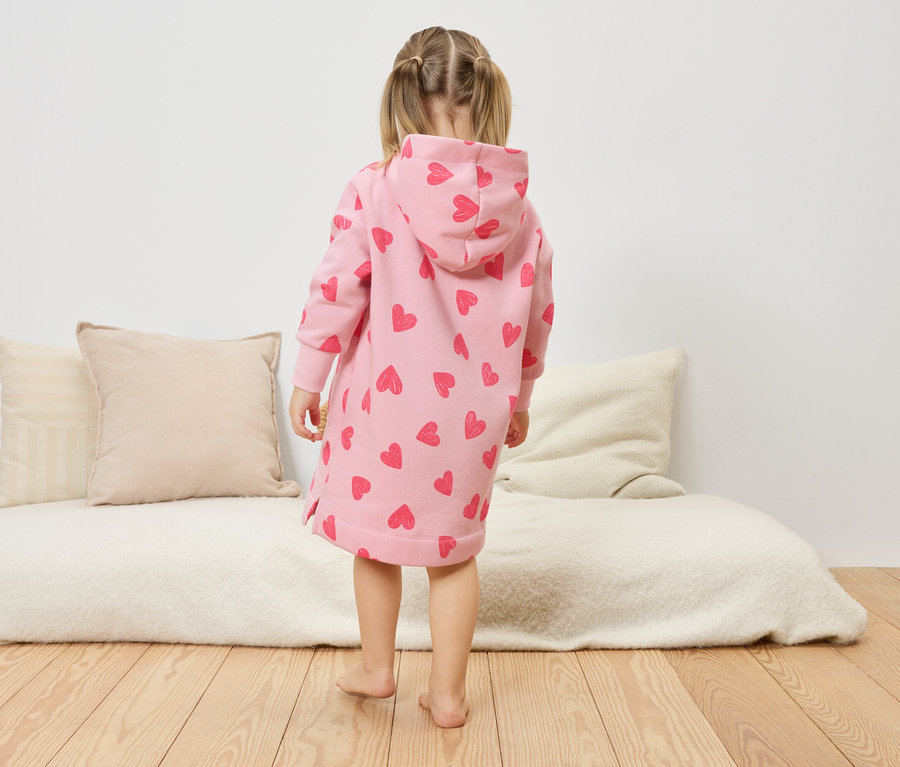 Holčička stojí zády ke kameře a má na sobě růžové šaty s kapucí a potiskem srdíček.