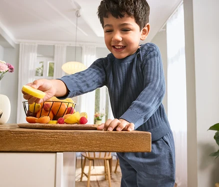 Chlapec v dětském pleteném svetru a dětských kalhotách drží plátek citronu nad stolem s ovocem.