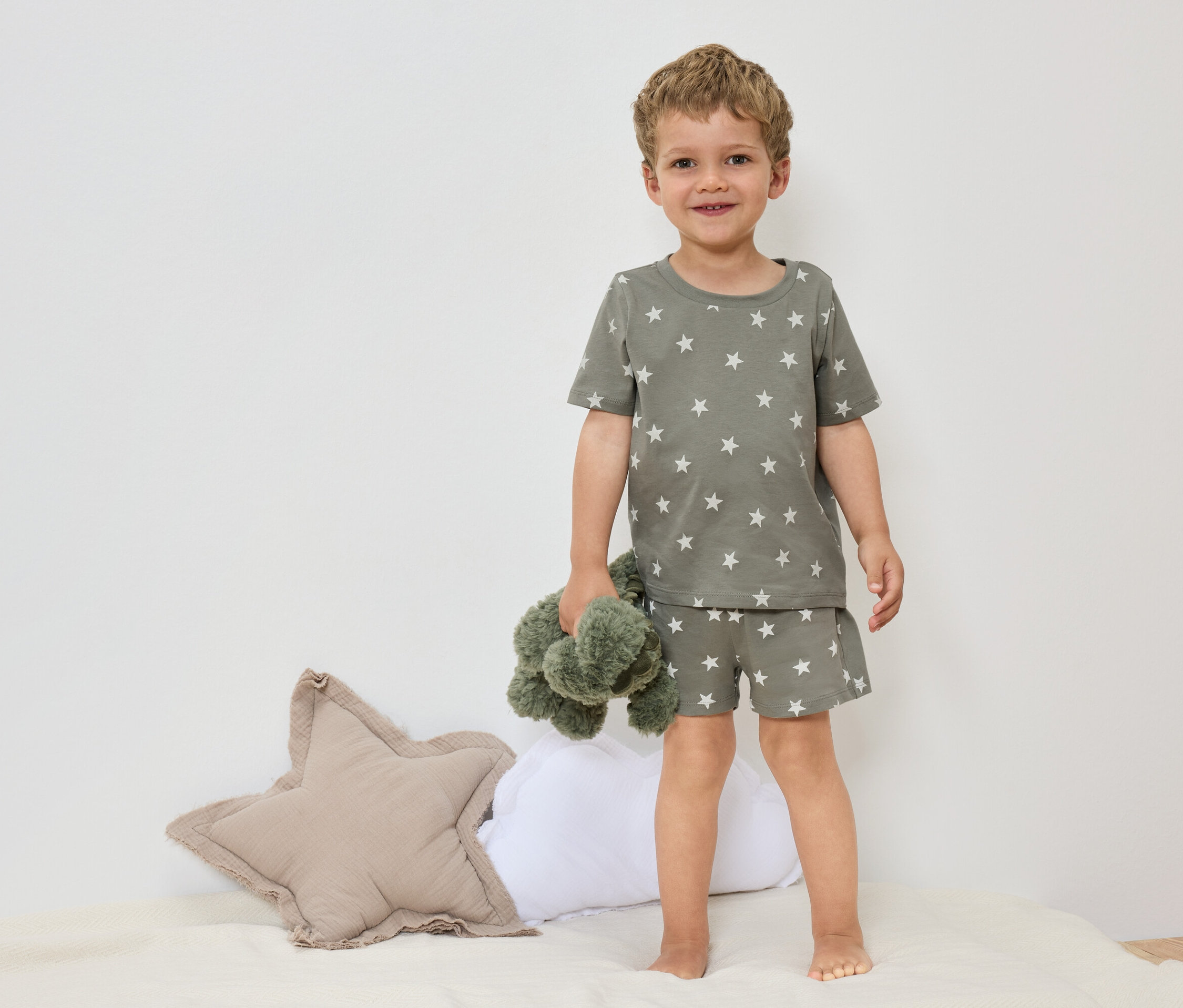 Malý chlapec stojí bosý na posteli v olivově zeleném pyžamu s hvězdami a drží zelenou plyšovou hračku.