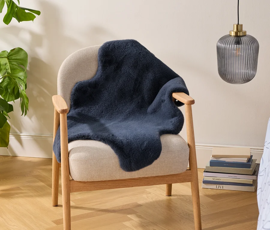 Béžové křeslo s tmavě modrým přehozem z umělé kožešiny, vedle něj velká monstera v proutěném koši. Na podlaze je stoh knih a nad ním visí žebrovaná skleněná lampa. Vpravo je vidět světle modré povlečení se vzorem.