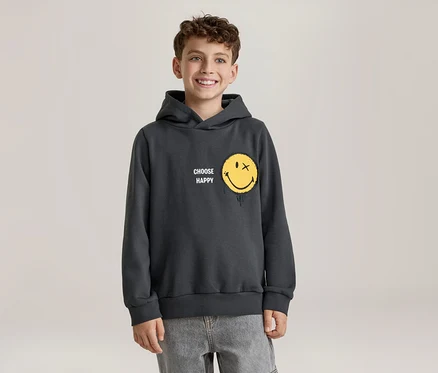 Usmívající se chlapec v tmavě šedé mikině s kapucí se žlutým smajlíkem, který má místo oka X a efekt tání, a nápisem „CHOOSE HAPPY“. Má na sobě světle šedé džíny.