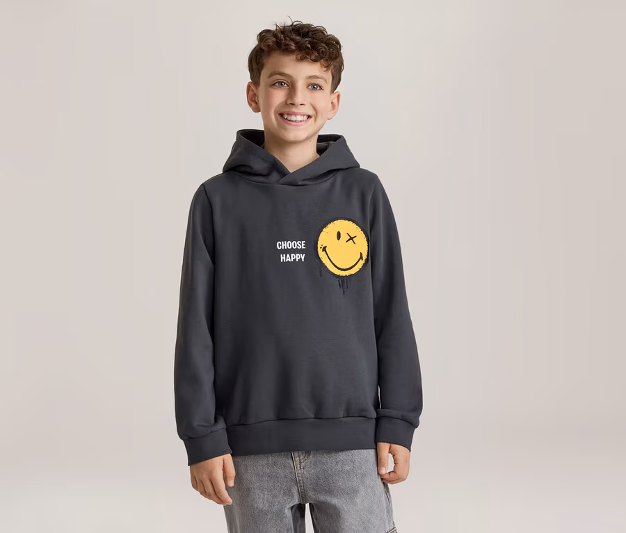 Usmívající se chlapec v tmavě šedé mikině s kapucí se žlutým smajlíkem, který má místo oka X a efekt tání, a nápisem „CHOOSE HAPPY“. Má na sobě světle šedé džíny.