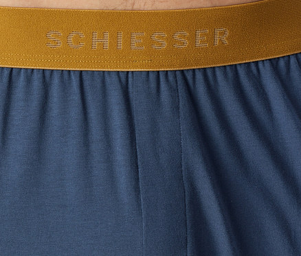 Detail modré látky se zlatým elastickým pasem, na kterém je perforovanými písmeny napsán název značky „SCHIESSER“.