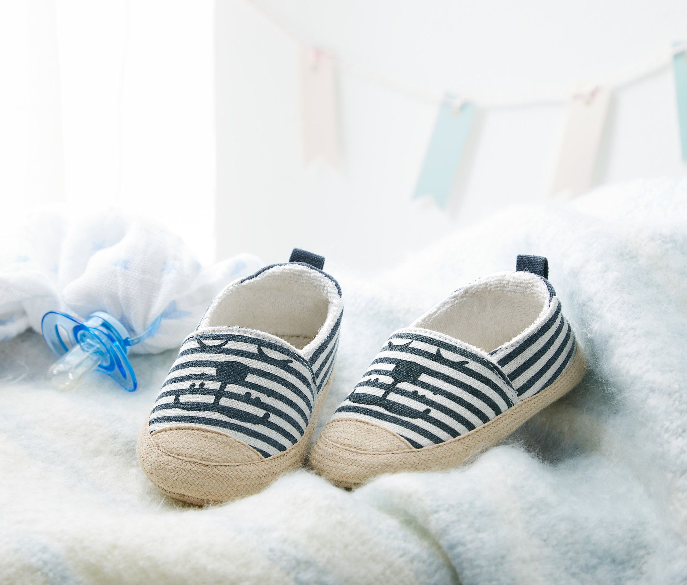 Dvoje boty pro batolata s modro-bílými pruhy leží na bílé dece vedle modrého dudlíku.
