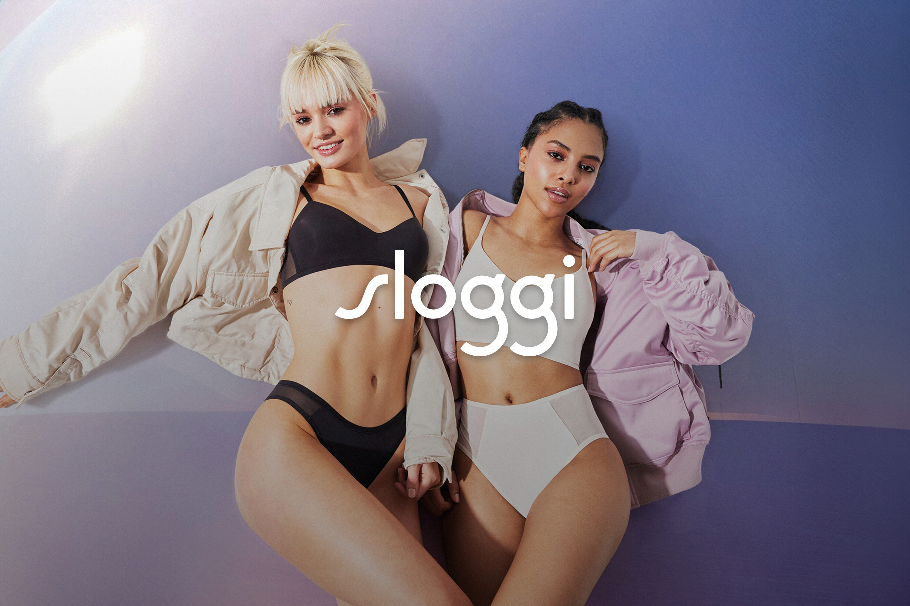 Dvě ženy pózují v černém a bílém spodním prádle s bundami před modro-fialovým pozadím.