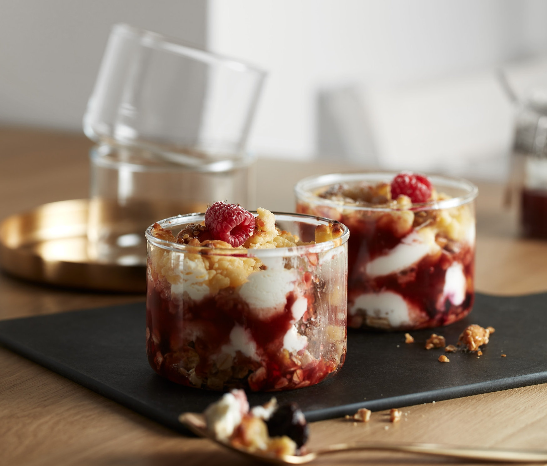 Na stole jsou 4 skleničky na dávkování a pečení s ovocným dezertem. Dvě skleničky jsou na černé podložce a dvě jsou naskládané na sobě na zlatém tácku.