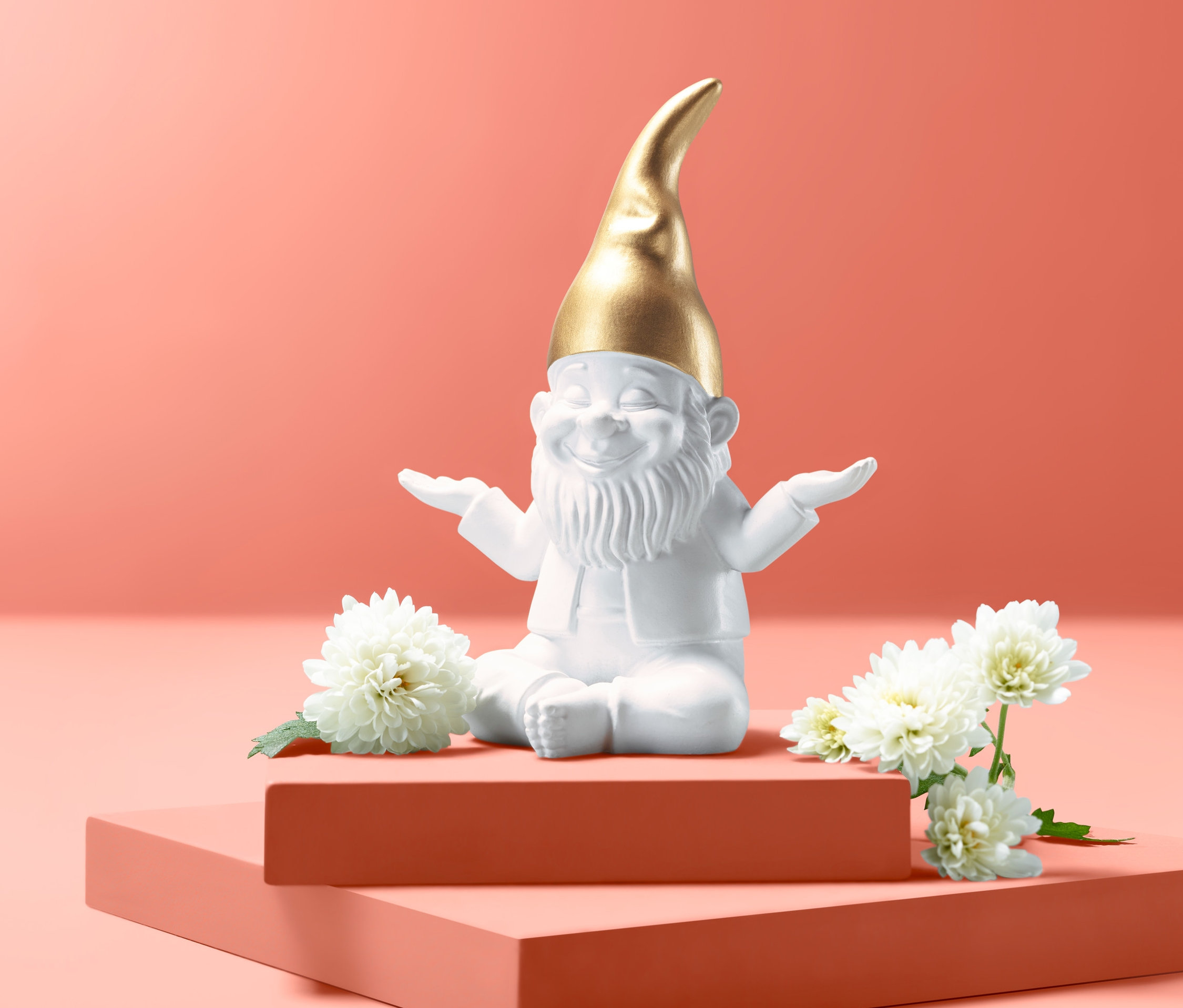 Bílý dekorační zahradní trpaslík se zlatou čepicí sedí na oranžovém podstavci a kolem něj jsou bílé květiny.