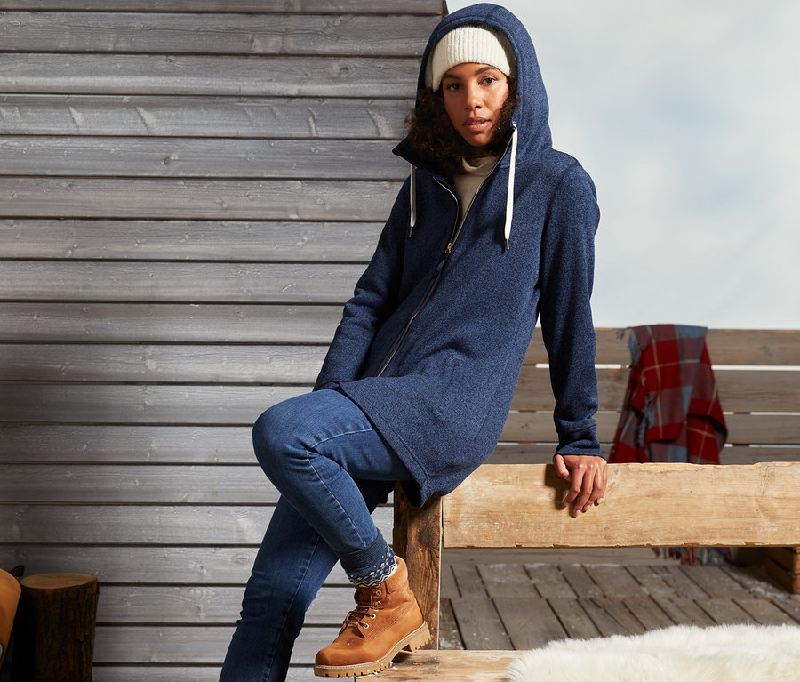 Žena sedí na dřevěné lavici a má na sobě pletený flísový kabát.