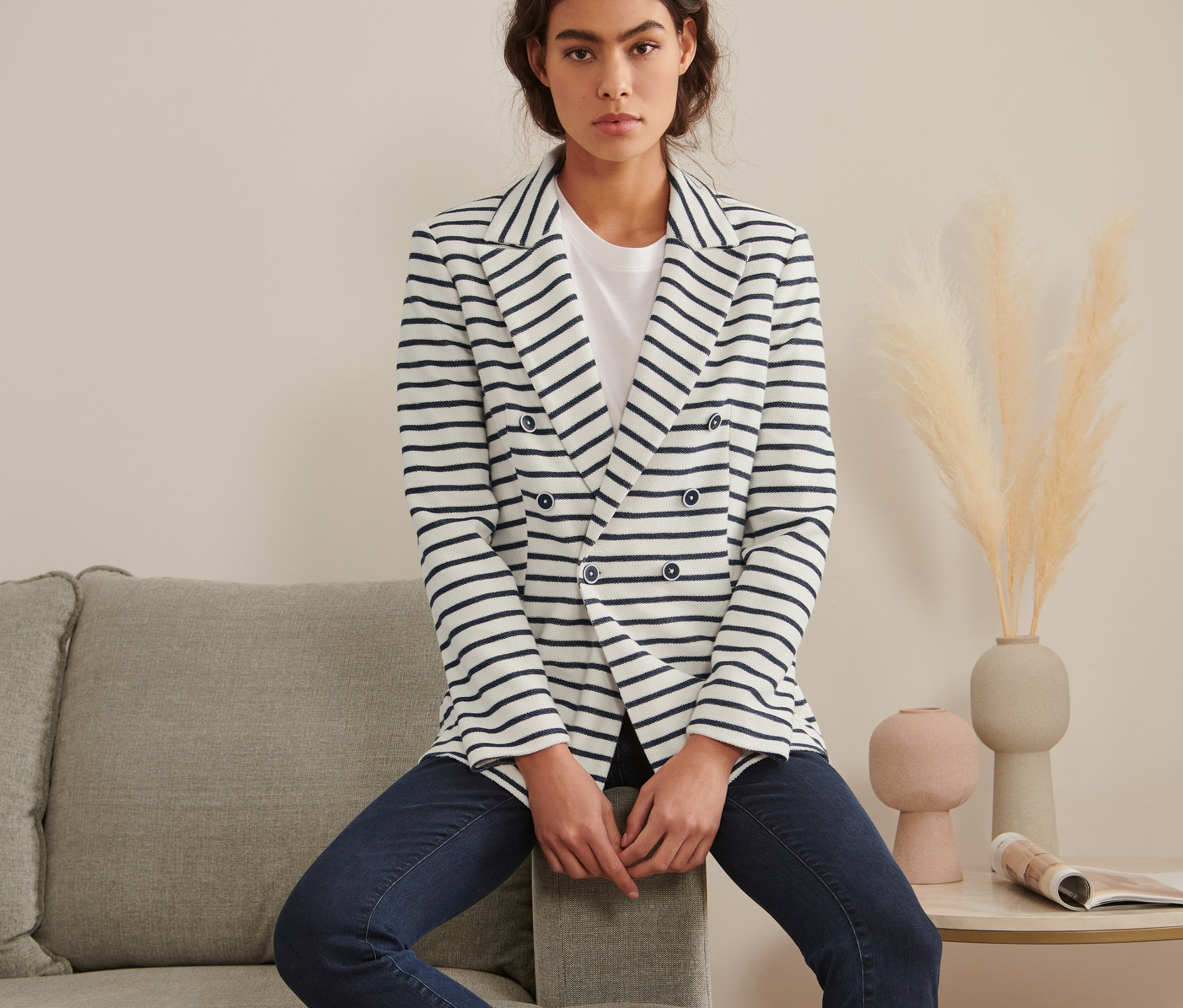Žena sedí na šedé pohovce a má na sobě proužkované sako.