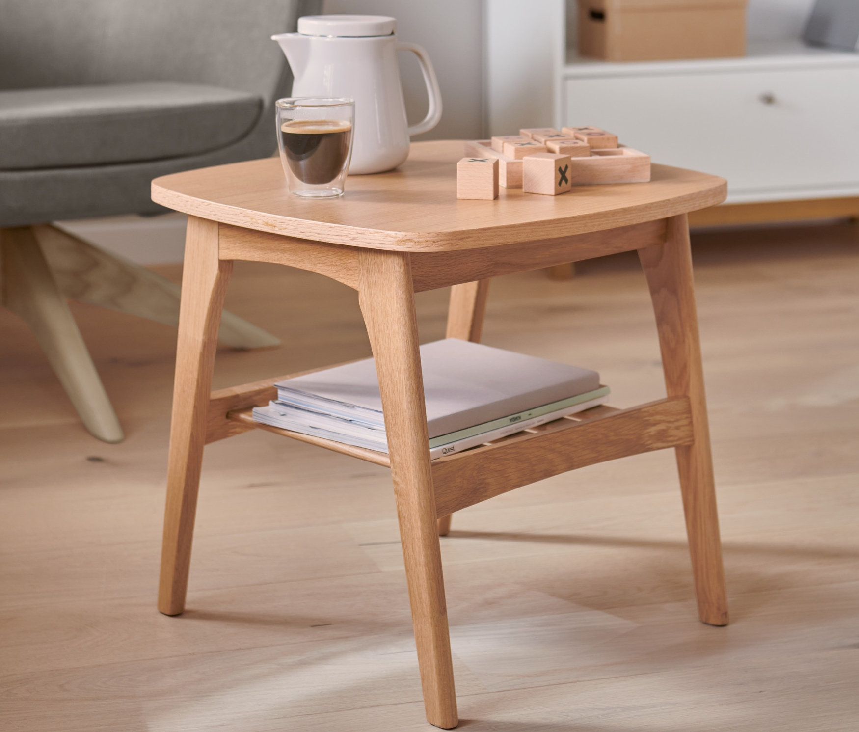 Na dřevěném stolku stojí kávovar, sklenice s kávou a dřevěné kostky. V pozadí je vidět šedé křeslo a bílá knihovna.