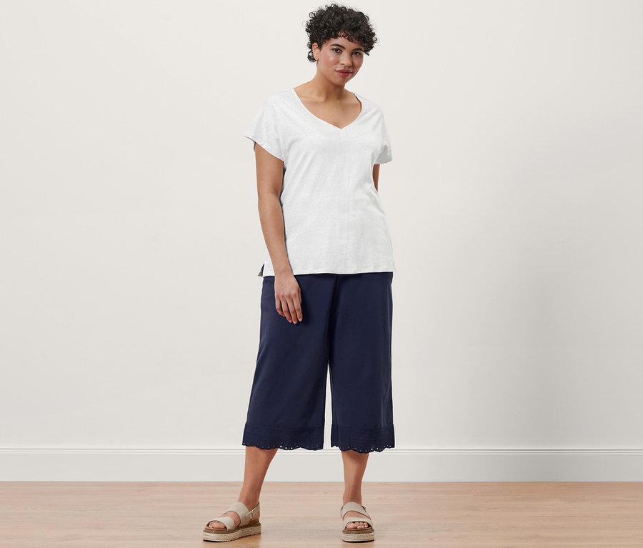 Žena pózuje v krémově bílém tričku ze směsi lnu, v tmavě modrých kalhotách culotte s výšivkou a kožených sandálech na platformě.