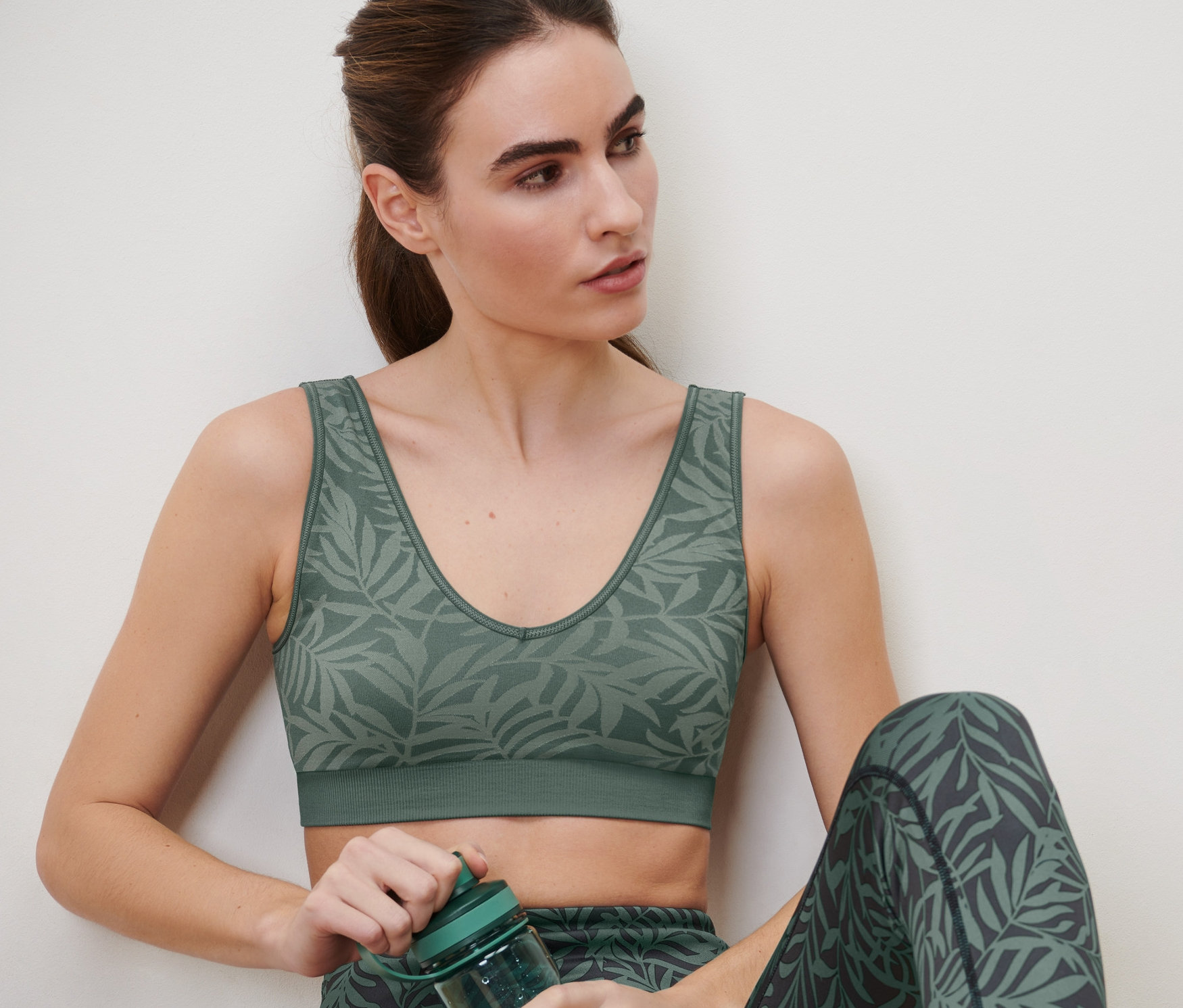Žena sedí a drží zelenou sportovní láhev. Má na sobě bezešvou sportovní podprsenku v šalvějové zelené barvě a legíny s potiskem listů.