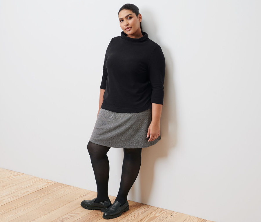 Žena opřená o zeď má na sobě černou mikinu se 3/4 rukávy, teplákovou sukni, černé punčochy a kožené mokasíny.