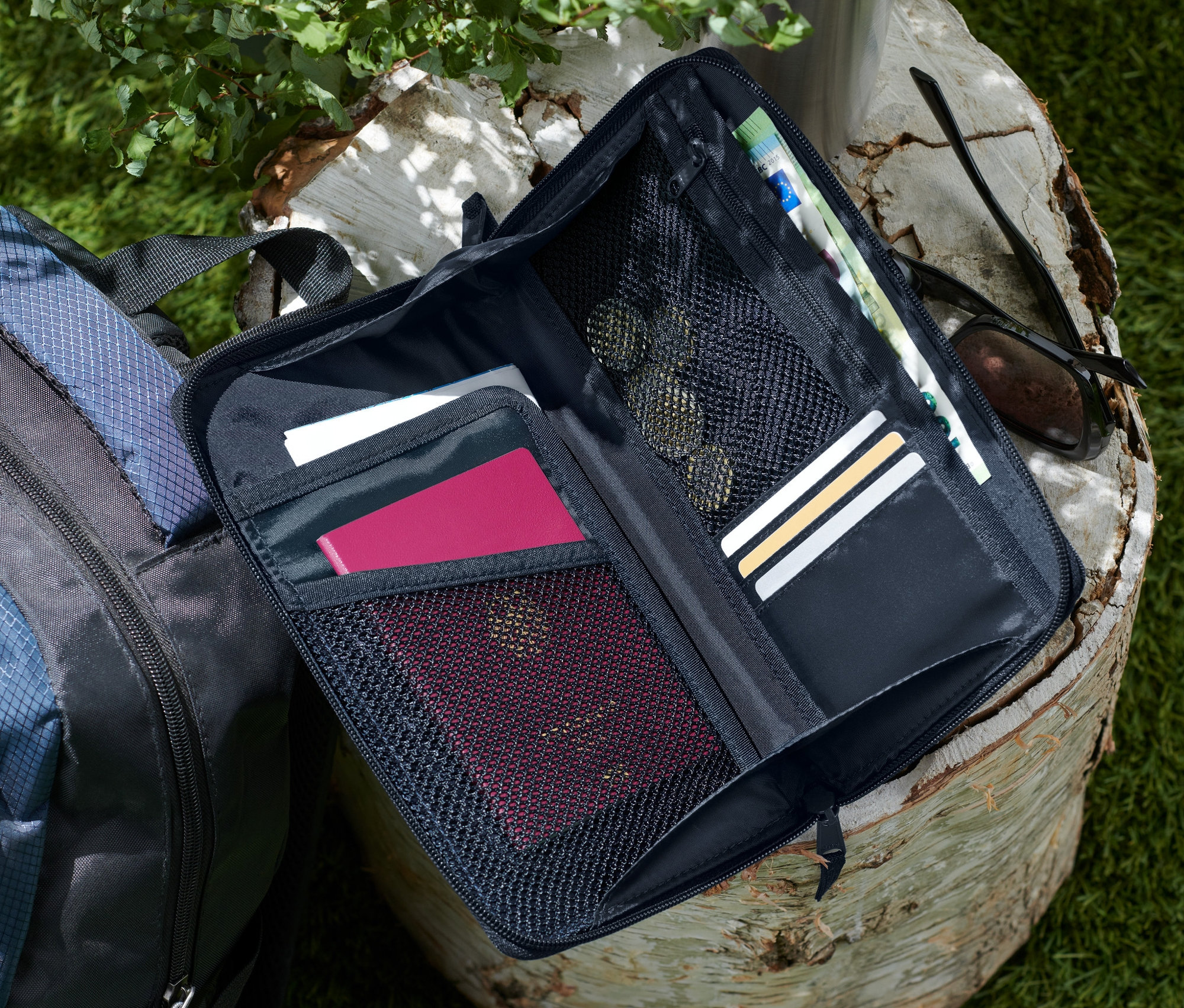 Otevřené černé pouzdro na doklady leží na pařezu vedle modro-černého batohu.