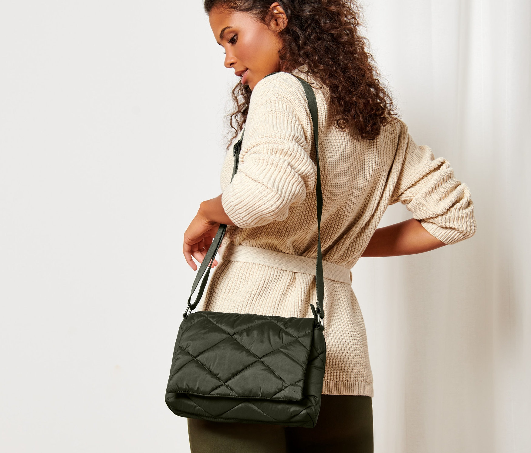 Žena s hnědými kudrnatými vlasy a v béžovém svetru s páskem pózuje s prošívanou kabelkou přes rameno.