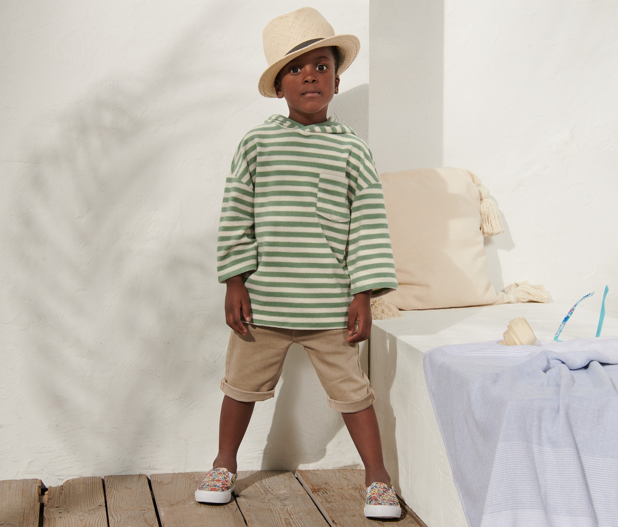 Chlapec v dětském froté tričku, béžových šortkách a klobouku stojí u bílé zdi.