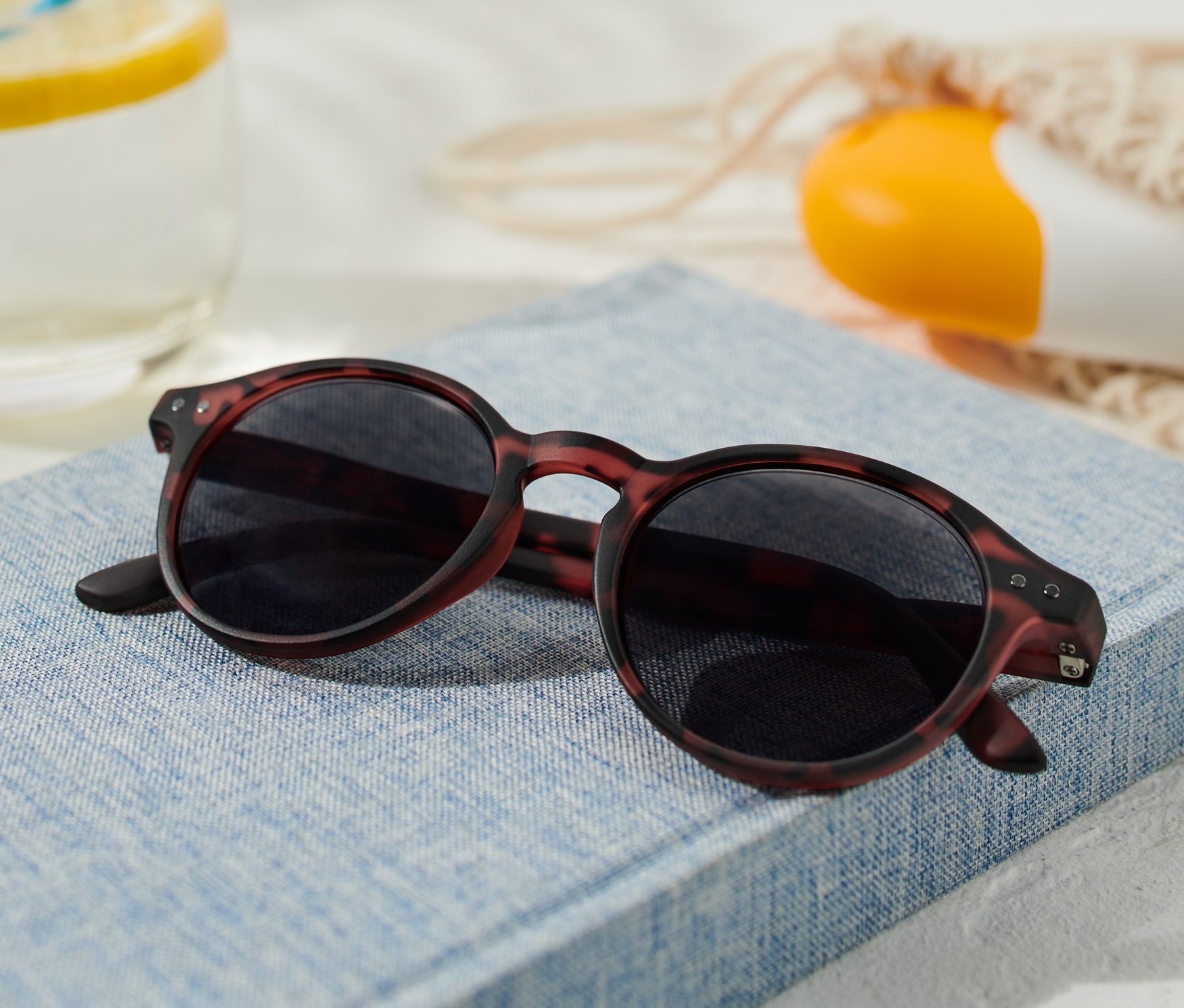 Sluneční brýle leží na modré knize vedle sklenice s citronem a opalovacího krému.