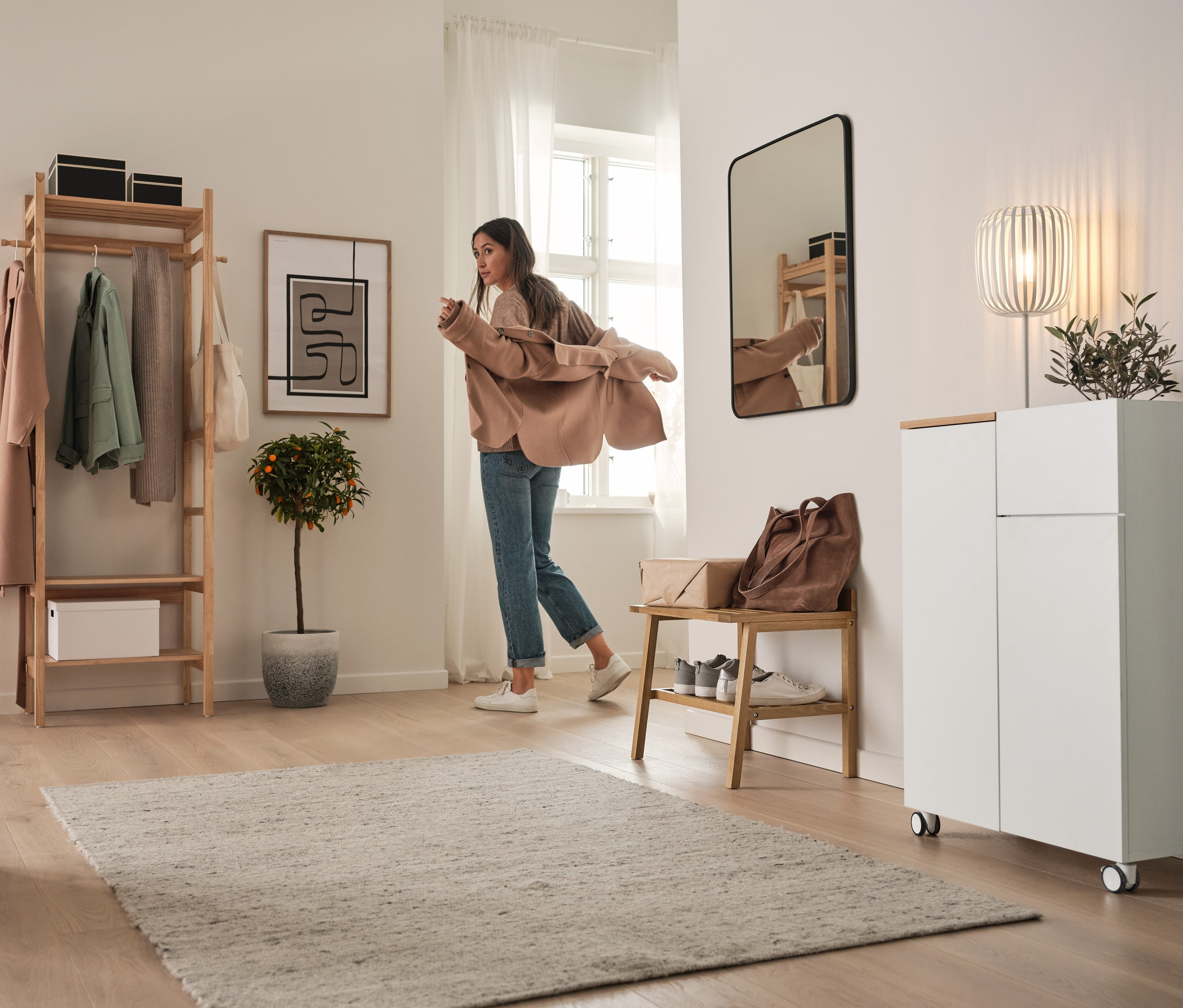 Žena si obléká sako v předsíni. V pozadí je otevřená šatní skříň s policemi, vysoká skříňka na kolečkách, kovová stolní lampa a stolička se širokými odkládacími plochami.