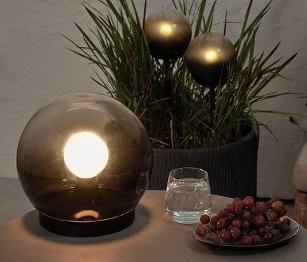 Na stolku stojí kulovitá lampa z kouřového skla, sklenice vody a hrozny. V pozadí jsou v květináči 2 LED kuličková světla.