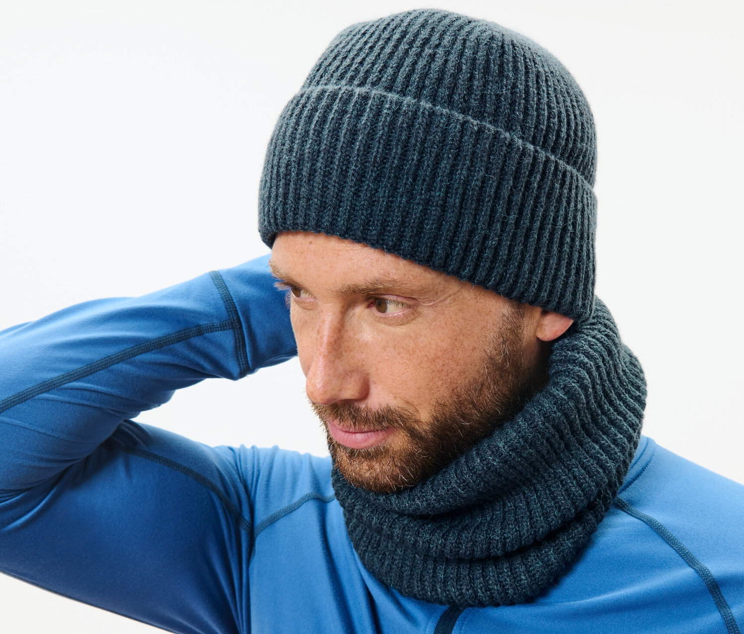 Muž v modrém svetru a pletené kruhové šále si upravuje pletenou čepici.