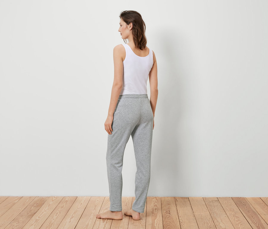 Žena stojí zády k divákovi a má na sobě bílé tílko a šedé tepláky.