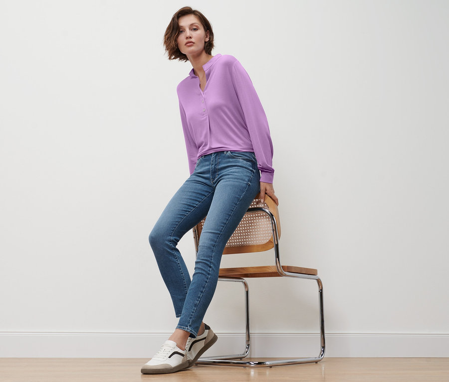 Žena sedí na židli a má na sobě džíny slim »Fit Emma«, středně modrý denim, šeříkové halenkové triko s knoflíkovou légou a kožené tenisky.